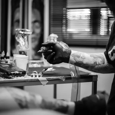 Tatoueur ganté tenant sa machine devant son poste de travail, en noir et blanc